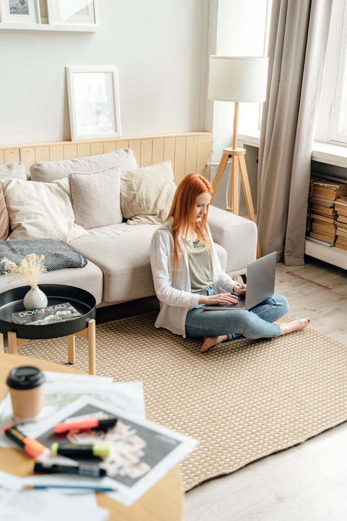 LES STUDIOS Young woman seated on a carpeted floor typing on a laptop in a cozy, sunlit living room.