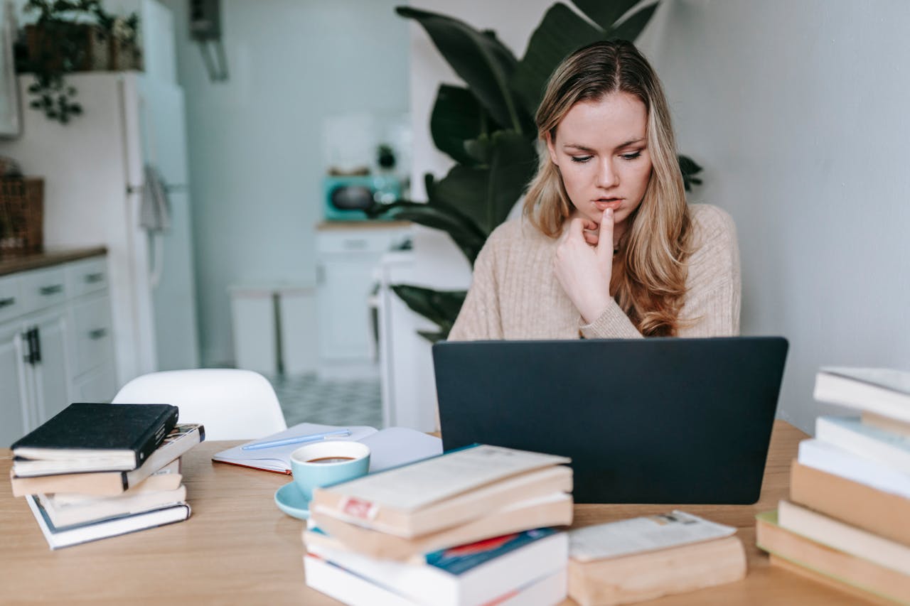 Contact A young woman studying at home, focused on her laptop, surrounded by books, symbolizing education and remote learning.